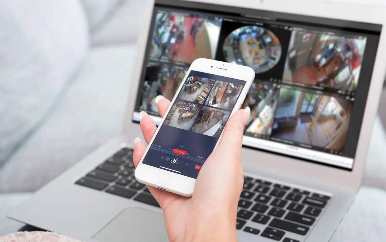 A hand holding a smartphone in front of a laptop. The laptop in the background is out of focus. The phone is in focus and shows the view from surveillance cameras. 